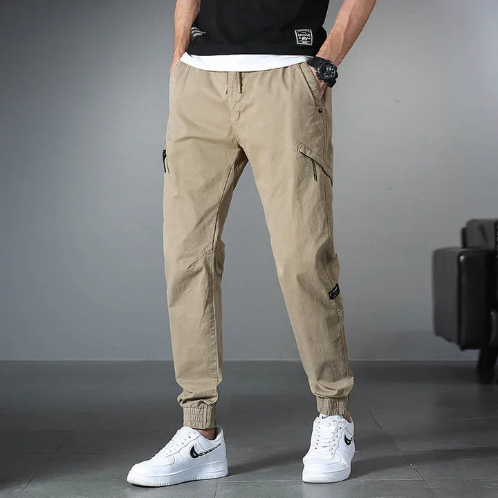 Men's beige cargo jogger pants with elastic cuffs paired with white Nike sneakers indoors