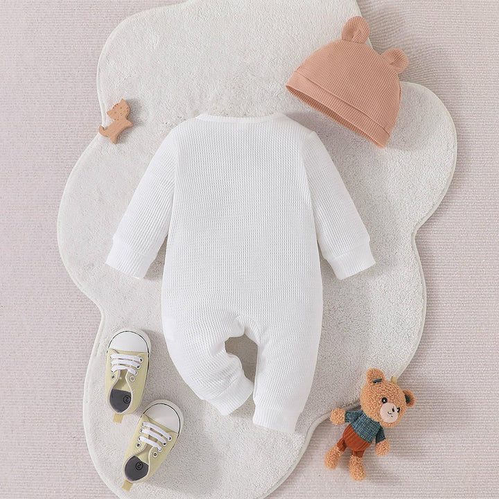 White textured baby onesie laid on a soft flower-shaped mat with brown bear hat, yellow baby shoes, and teddy bear toy
