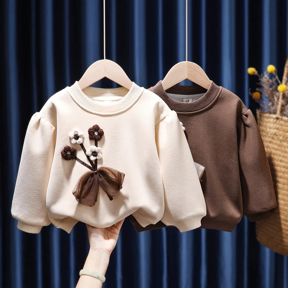 Two children's sweatshirts on wooden hangers, one cream with floral applique and bow, one solid brown
