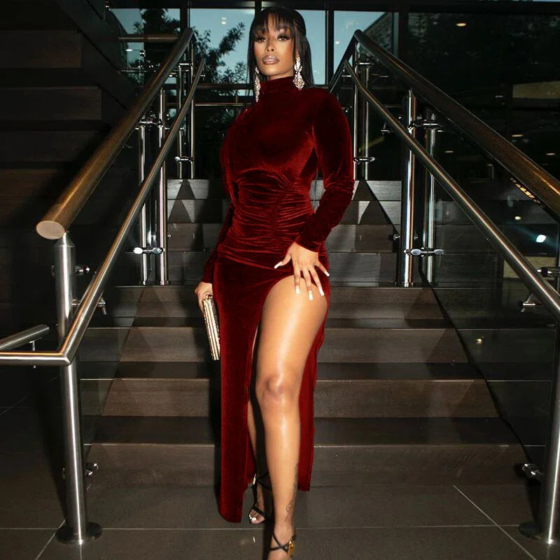 Woman wearing a high-slit burgundy velvet evening dress with long sleeves and holding a gold clutch on stairs