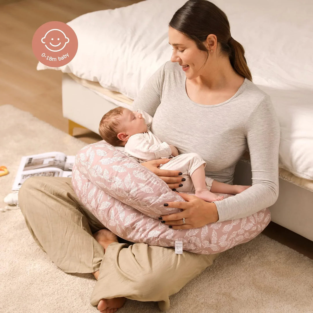 Mother sitting on carpeted floor holding baby in pink patterned nursing pillow in bedroom