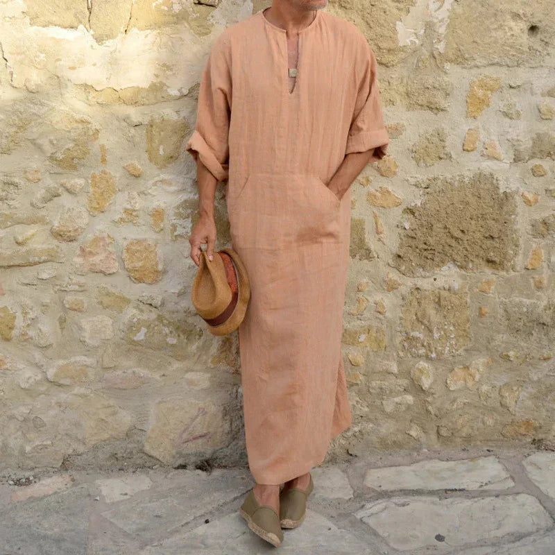 Man wearing beige long linen robe and holding straw hat standing against stone wall