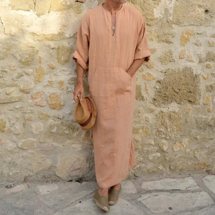 Man wearing beige long linen robe and holding straw hat standing against stone wall