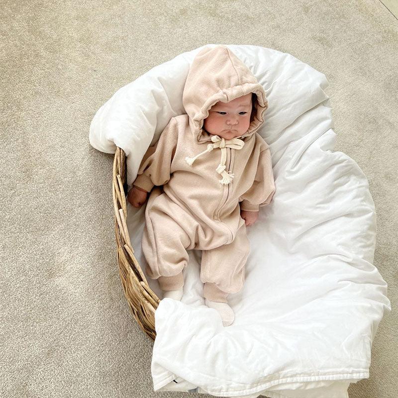 Baby in beige hooded onesie lying in wicker basket with white blanket on beige carpet