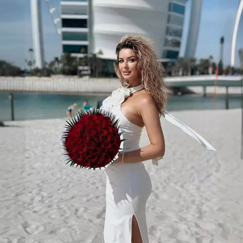 Woman in white dress holding large bouquet of red roses on sandy beach with modern building backdrop