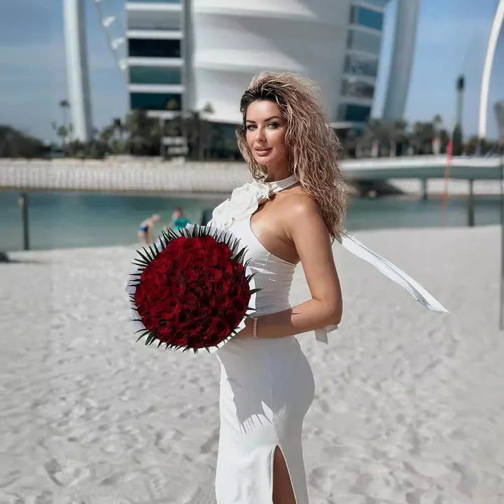 Woman in white dress holding large bouquet of red roses on sandy beach with modern building backdrop