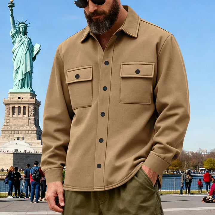 Bearded man wearing a tan button-up shirt with chest pockets near Statue of Liberty in sunny outdoor setting