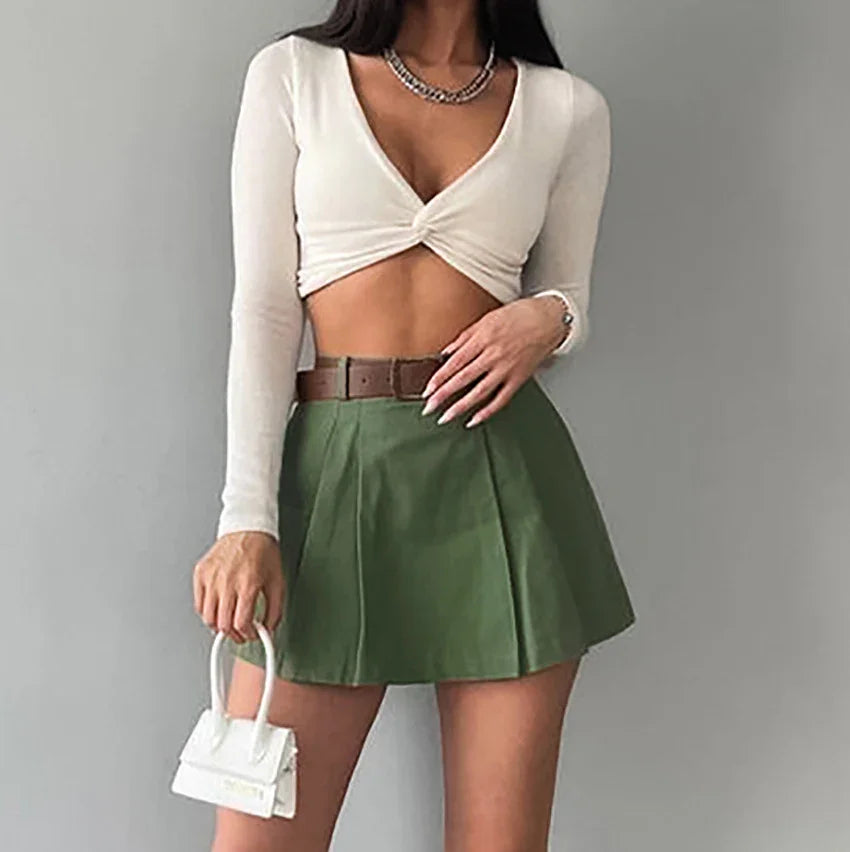 Woman wearing white twisted crop top, green pleated mini skirt with brown belt, holding small white handbag