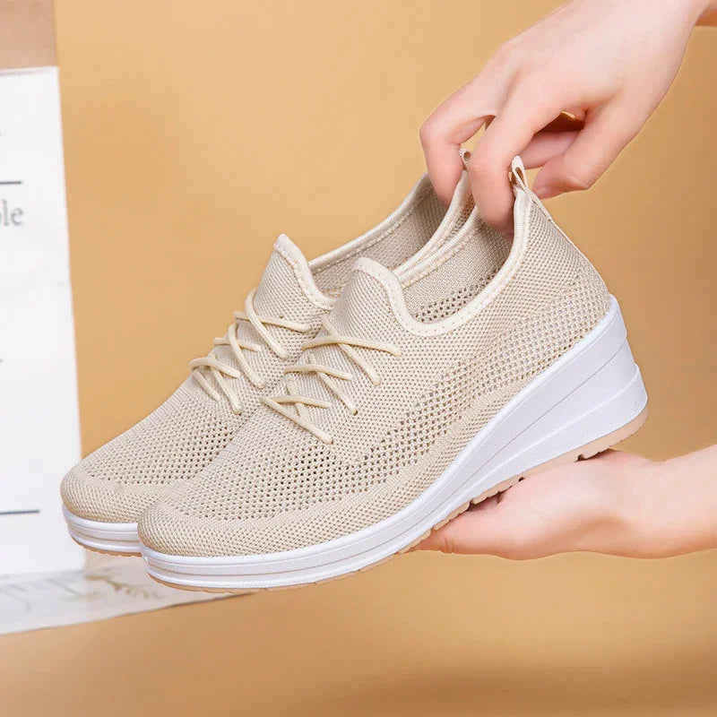 Hands holding beige knit casual sneakers with white wedge soles on tan background