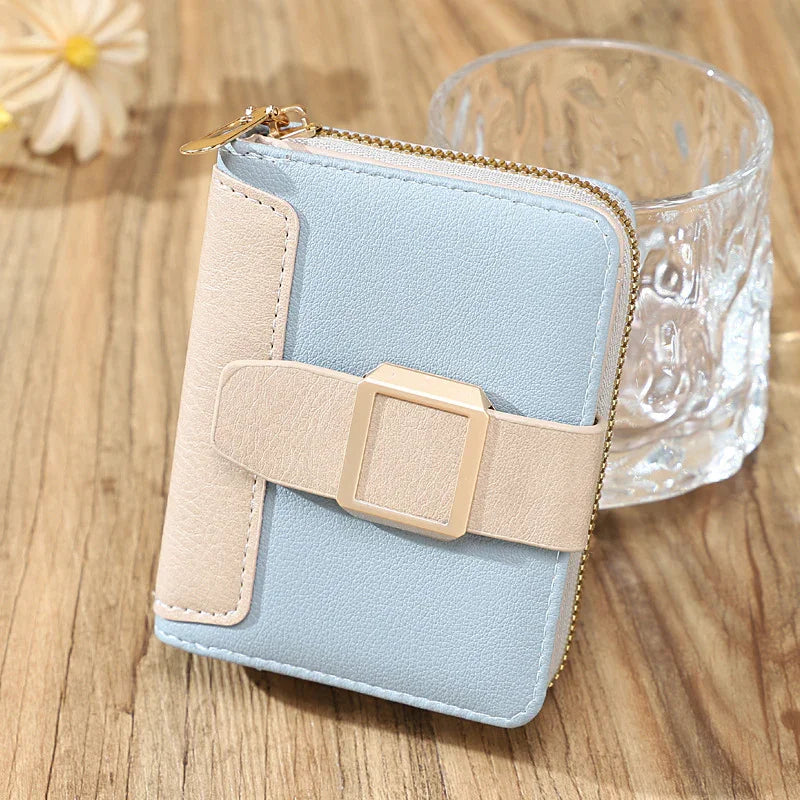 Light blue and beige leather zippered wallet with gold buckle on wooden table next to glass and flower