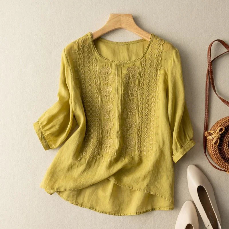 Yellow linen blouse with floral embroidery, 3/4 sleeves, round neckline laid flat with white shoes and a woven bag