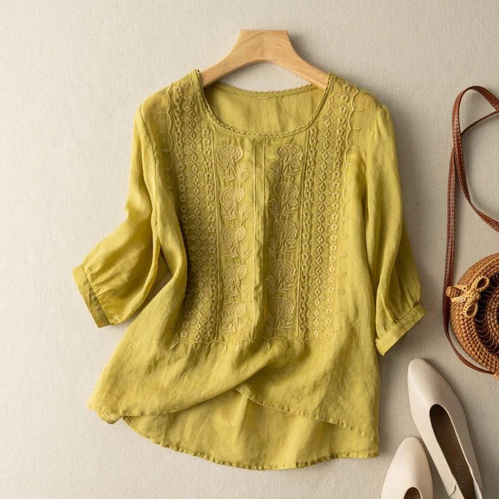 Yellow linen blouse with floral embroidery, 3/4 sleeves, round neckline laid flat with white shoes and a woven bag