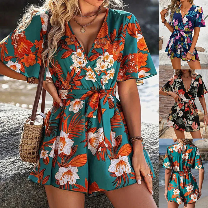 Woman wearing teal floral print romper with short sleeves and belt, carrying woven handbag by water