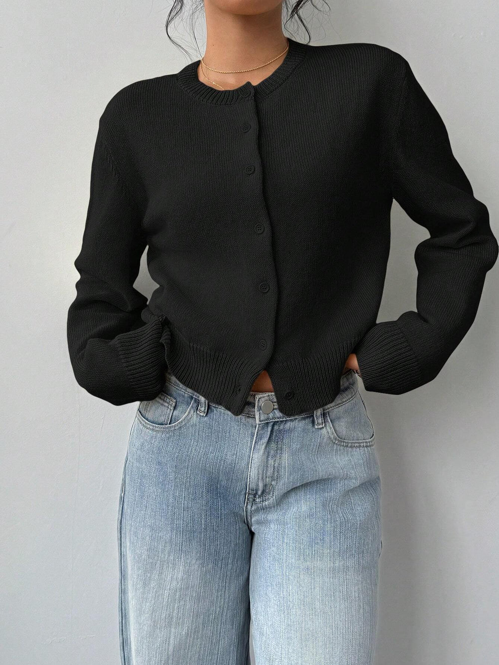Woman wearing black buttoned knit cardigan and light blue high-waisted jeans against plain wall