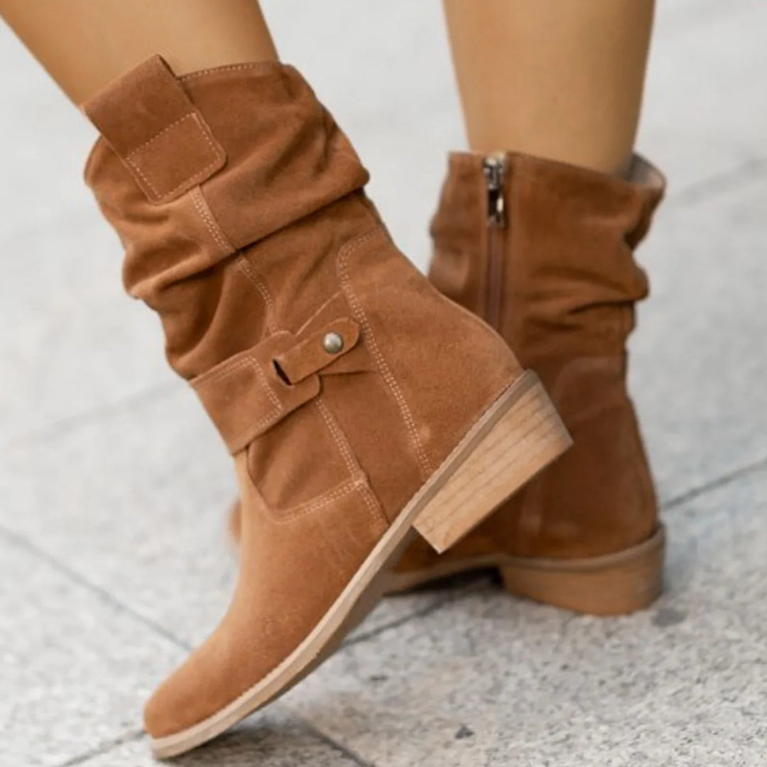 Tan suede ankle boots with slouchy design, side buckle strap, and low stacked heel on pavement
