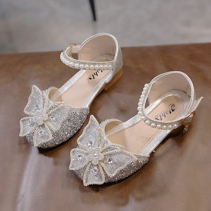 Sparkly silver girls' dress shoes with pearl ankle straps and decorative rhinestone bows on brown surface