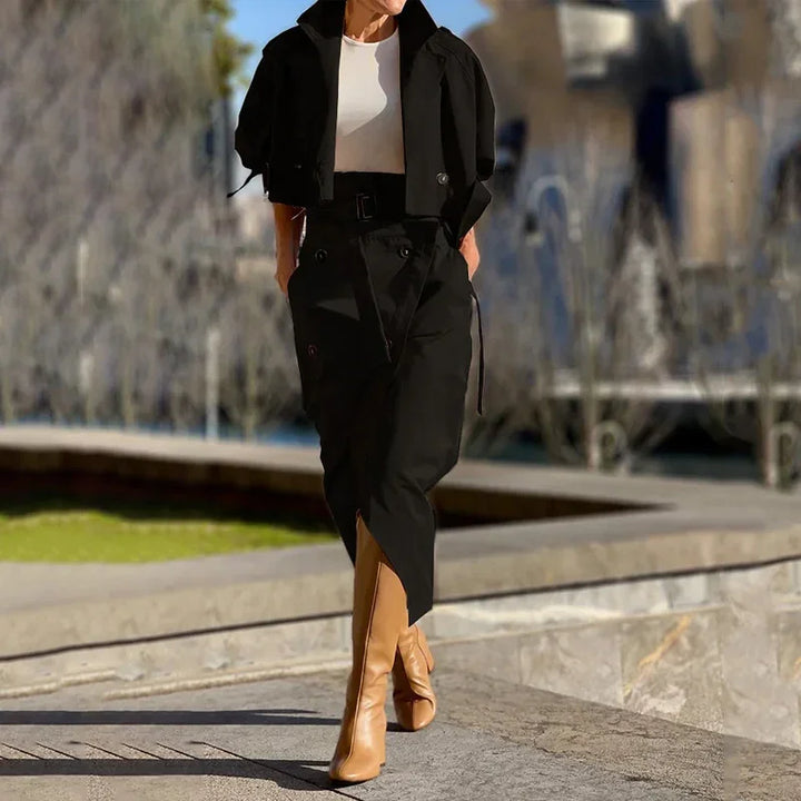 Woman wearing black belted skirt, cropped black jacket, white top, and tan knee-high boots outdoors
