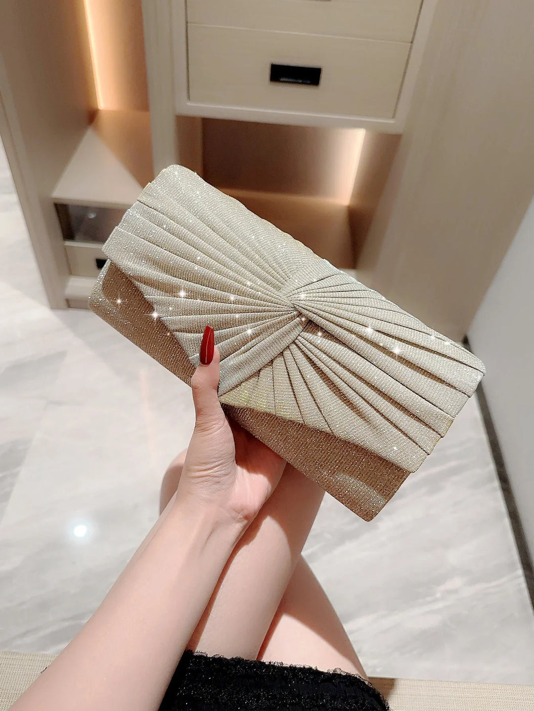 Hand holding a sparkly gold pleated clutch purse with red nails in a modern room