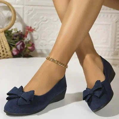 Navy blue wedge shoes with large bows worn on feet, paired with gold anklet, indoor setting