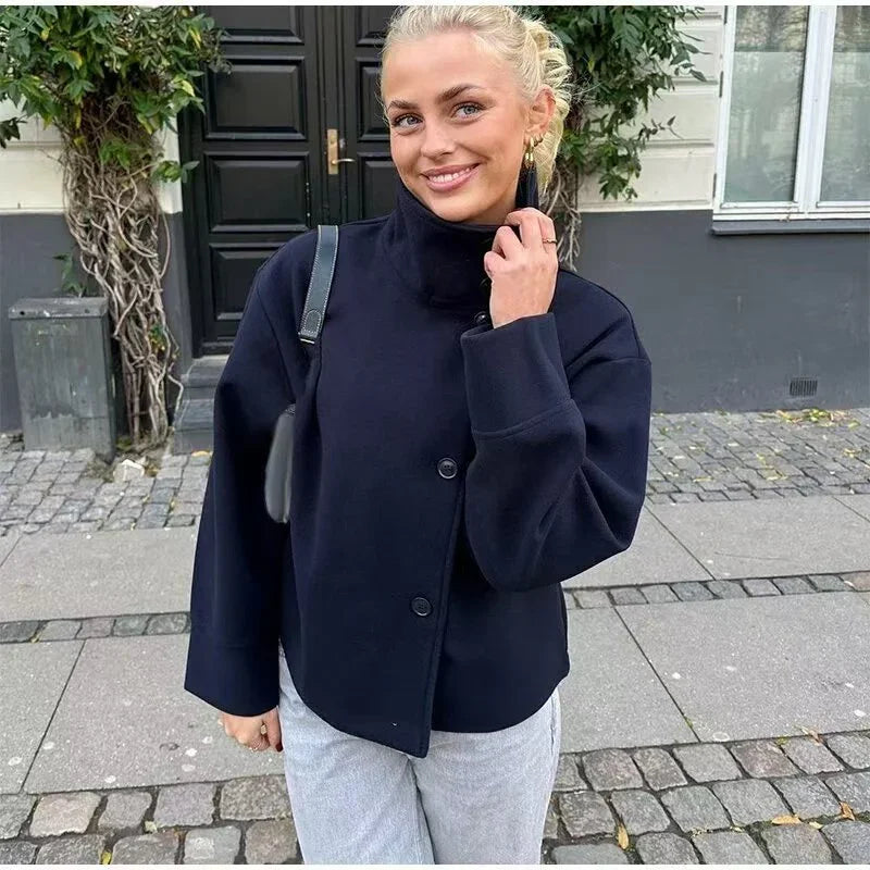 Smiling woman wearing a black buttoned jacket and light gray pants standing on a cobblestone sidewalk