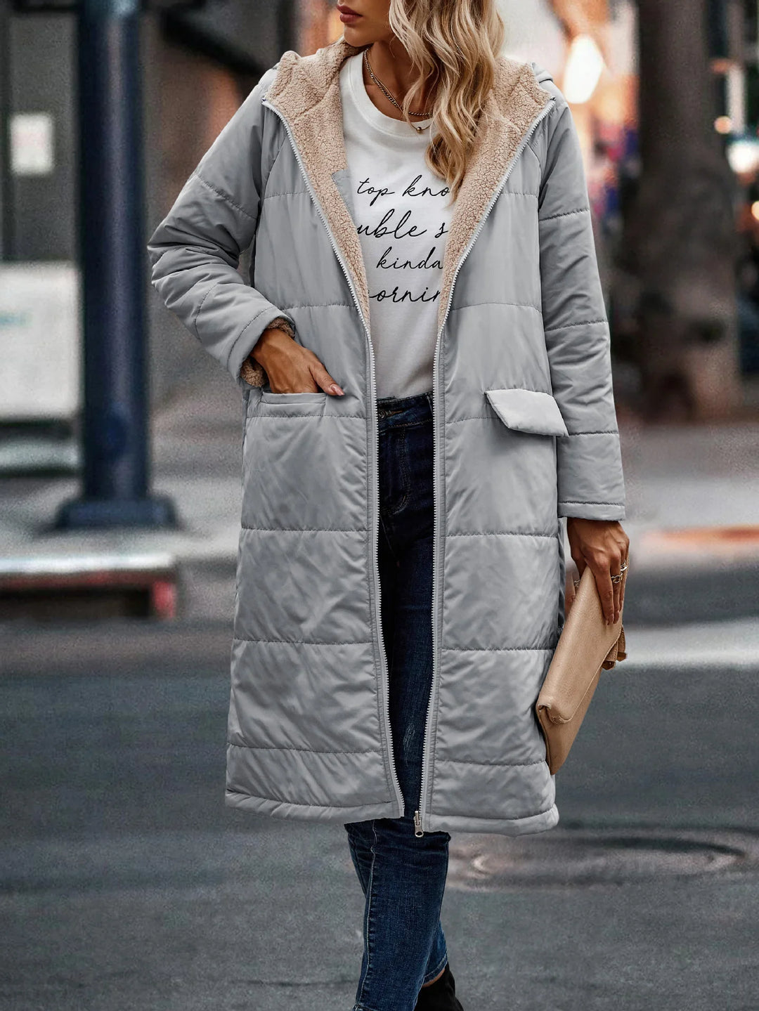 Woman wearing a light gray long padded winter coat with beige fleece lining, paired with dark jeans and a white printed T-shirt