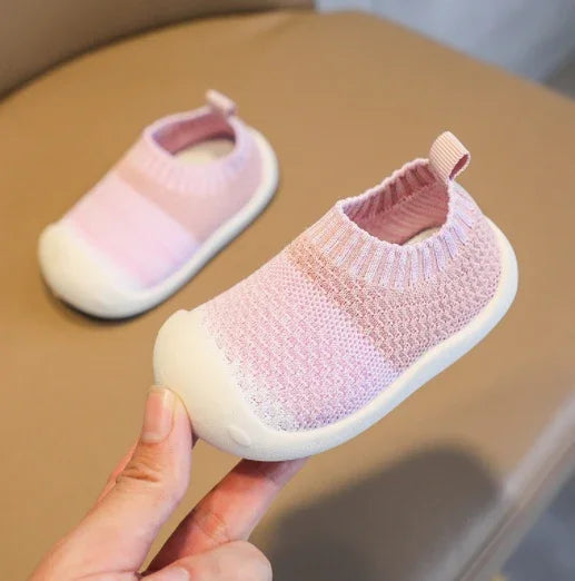 Light pink baby shoes with knit texture and white rubber soles on beige surface