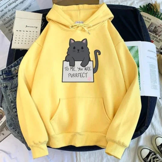 Yellow hoodie with a cute black cat holding a sign that reads 'To me, you are purrfect' laid on denim and black jeans