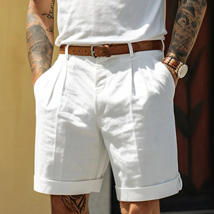 Man wearing white pleated shorts with rolled cuffs, brown belt, white shirt, tattoos on arms, and wristwatch