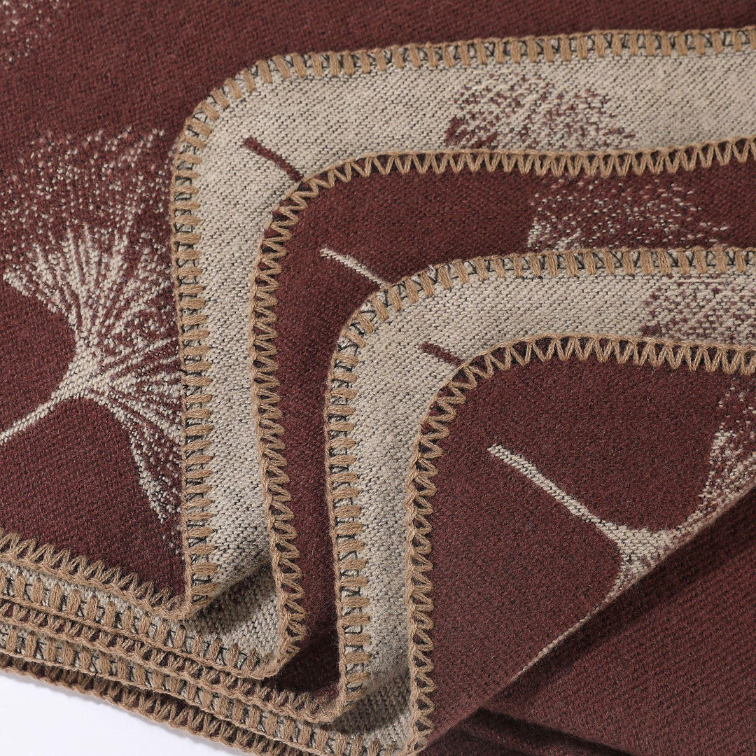 Close-up of a folded brown and beige woven blanket with botanical leaf patterns and stitched edges