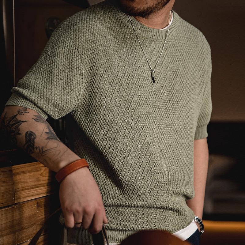Man wearing textured olive green short-sleeve sweater with silver lightning necklace and tattoos on arm