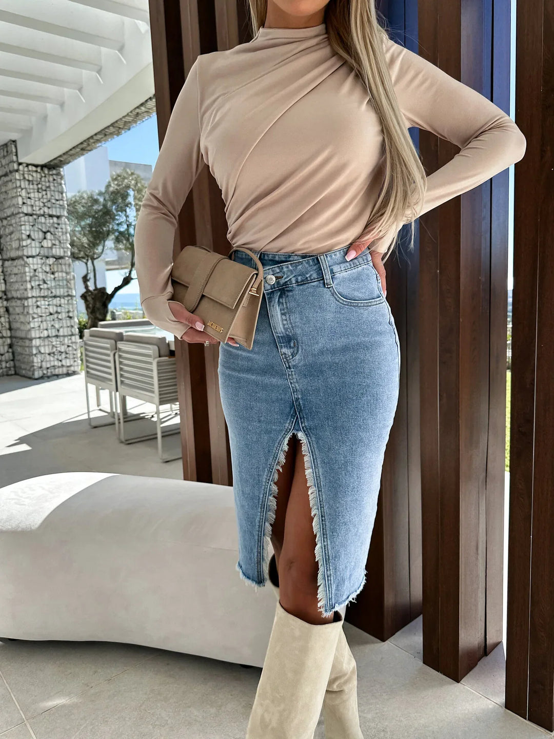 Woman wearing beige long-sleeve top, high-waisted denim skirt with front slit, beige boots, and holding beige purse