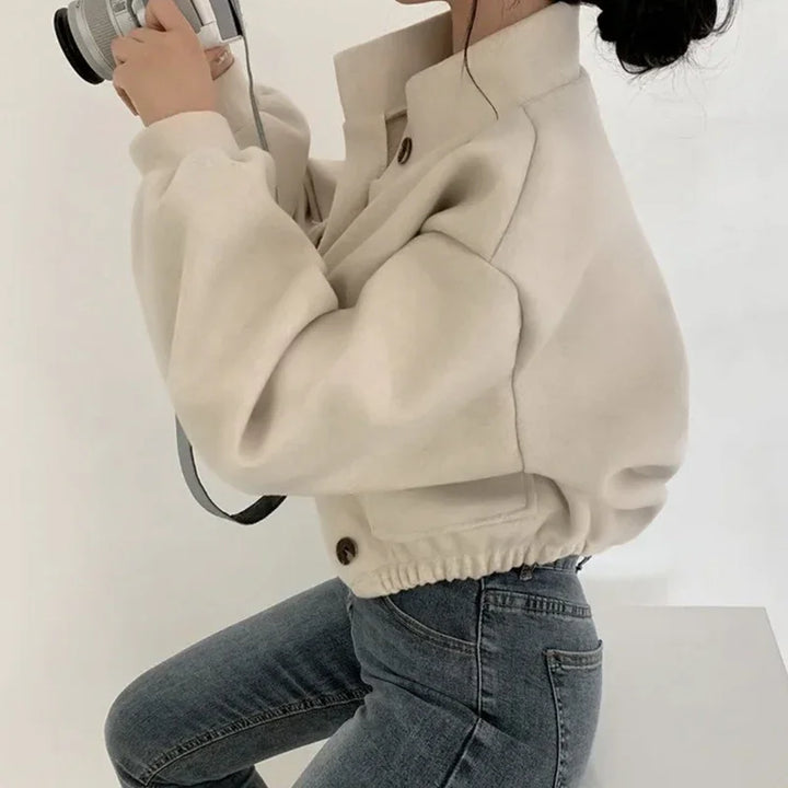 Woman in beige cropped jacket and blue jeans holding a camera in minimalist setting
