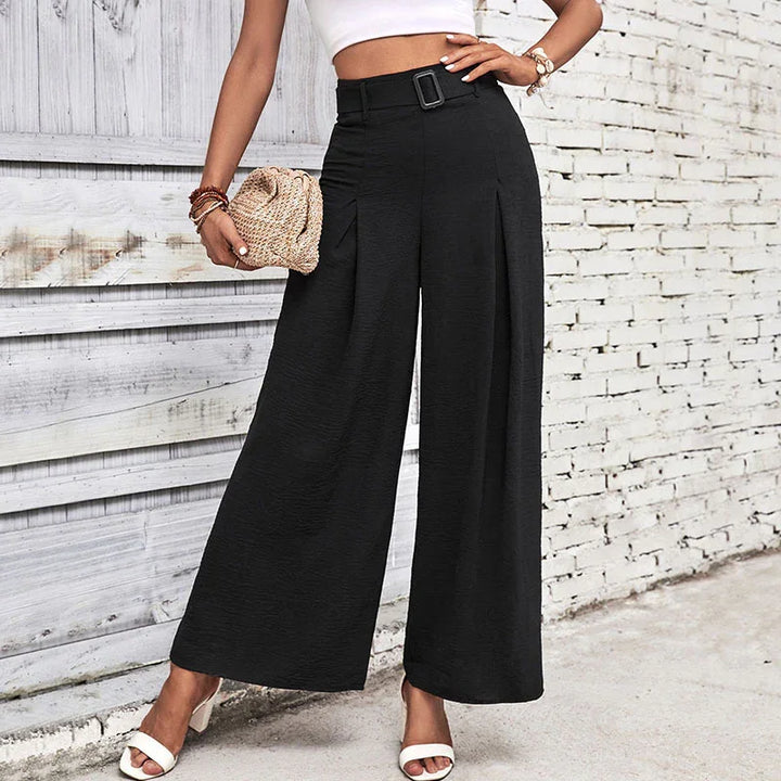 Woman wearing black wide-leg pants with belt and white strappy heels against white brick wall