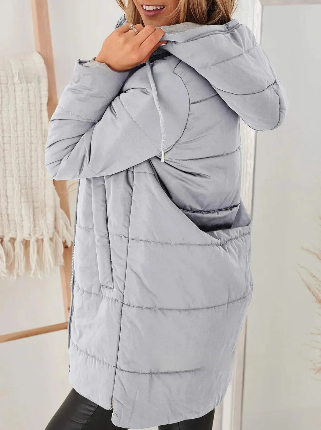 Woman wearing light gray puffer jacket with hood and pockets in cozy indoor setting