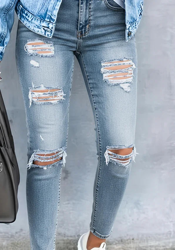 Close-up of legs wearing light blue ripped skinny jeans and denim jacket, standing against gray wall