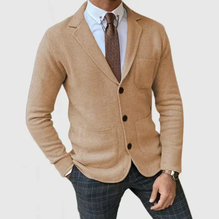 Man wearing beige knitted blazer with black buttons, white shirt, brown tie, and plaid pants