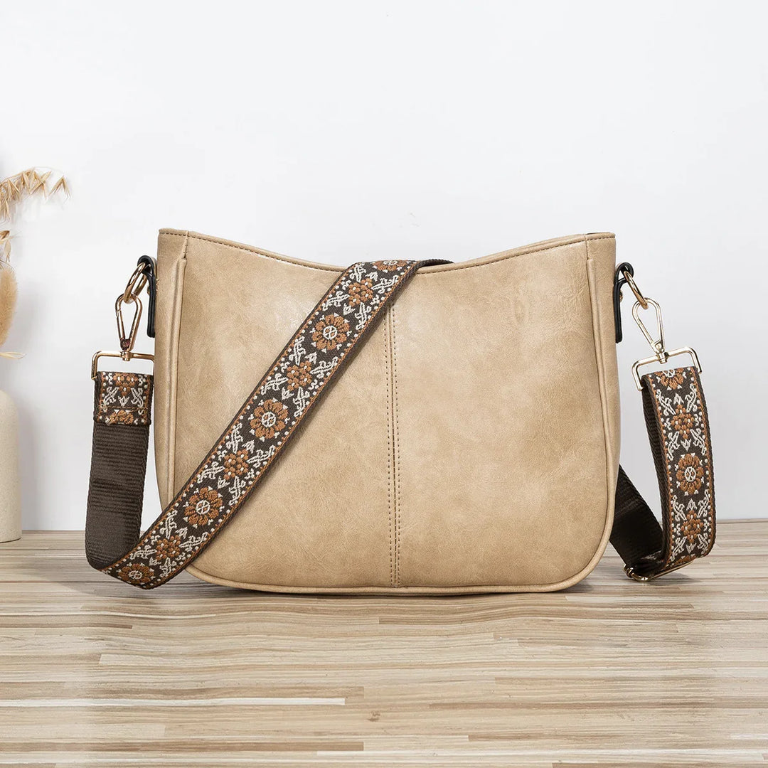 Beige leather crossbody bag with detachable embroidered floral strap on wooden surface