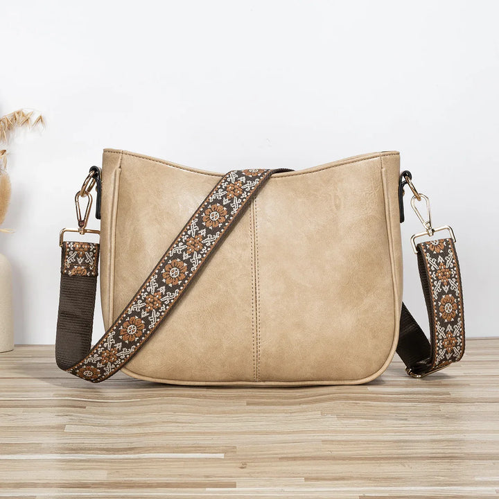 Beige leather crossbody bag with detachable embroidered floral strap on wooden surface