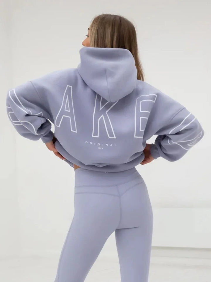 Woman wearing lavender hoodie with large white letters and matching leggings in minimal white room