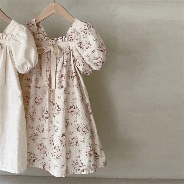 Two toddler dresses, one beige and one floral with puff sleeves, hanging on wooden hangers