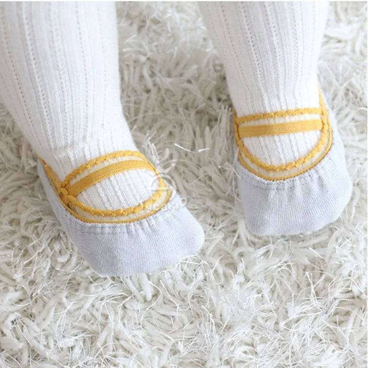 Baby feet wearing white socks with yellow decorative straps on a fluffy white rug