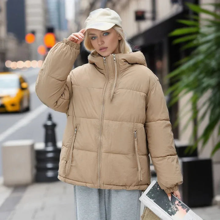 Blonde woman in beige puffer jacket and white cap holding a magazine on city street