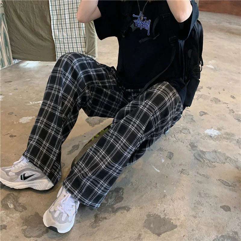 Person wearing black and white plaid pants, white sneakers, and a black t-shirt sitting on a concrete floor