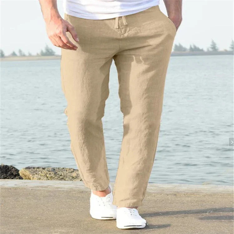 Man wearing beige linen pants and white sneakers walking near waterfront with trees in background