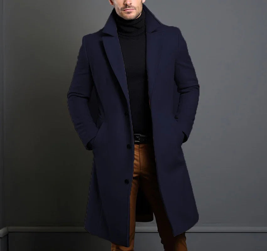 Man wearing navy blue overcoat and black turtleneck with brown pants on gray background