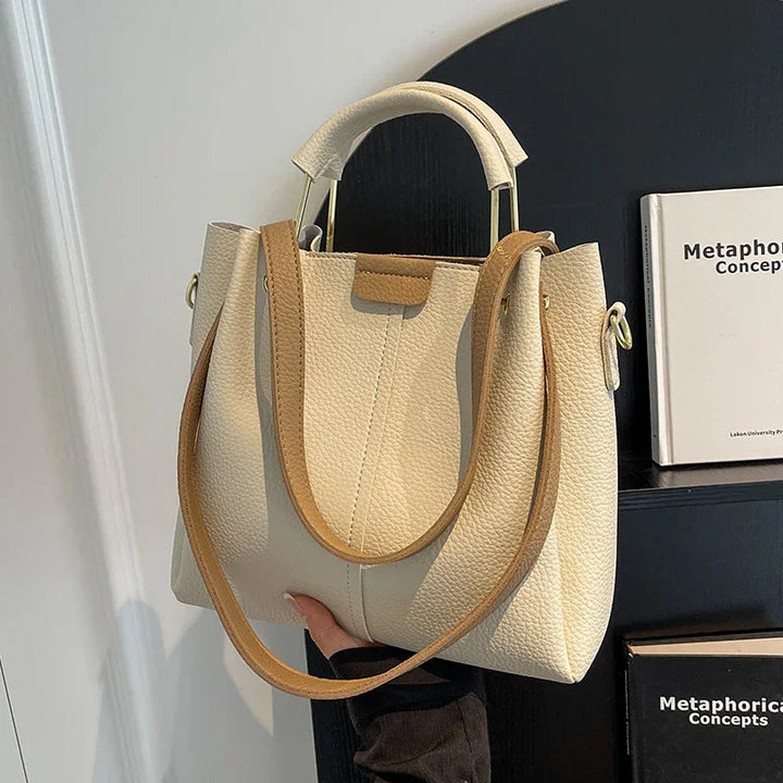 Cream textured leather handbag with tan strap and gold handles, held indoors by a person