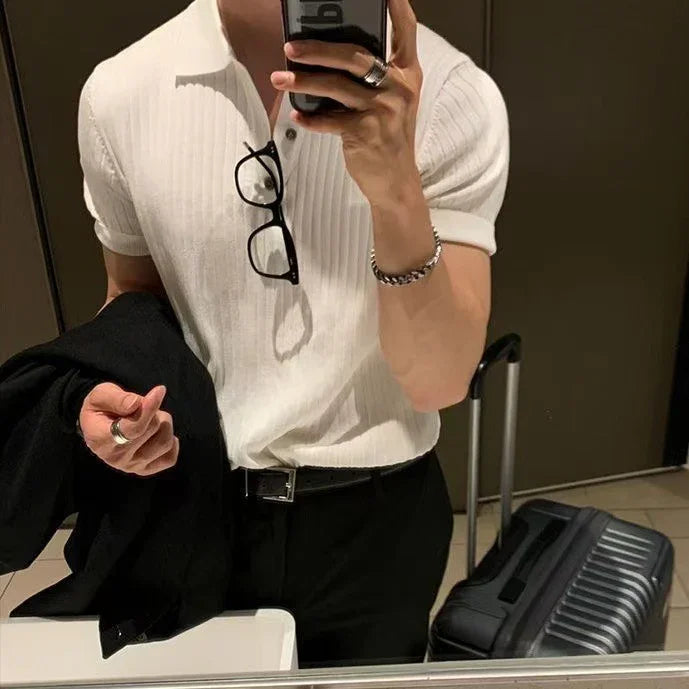 Man in white ribbed polo shirt with black glasses and accessories holding black jacket near a black suitcase in mirror selfie