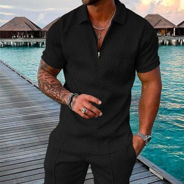 Man wearing black textured short-sleeve shirt and pants with tattooed arm on wooden pier by overwater bungalows