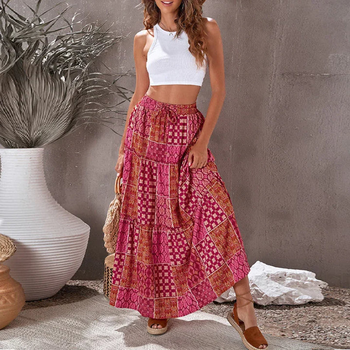 Woman wearing white crop top and long pink patterned skirt holding woven bag in minimalist room
