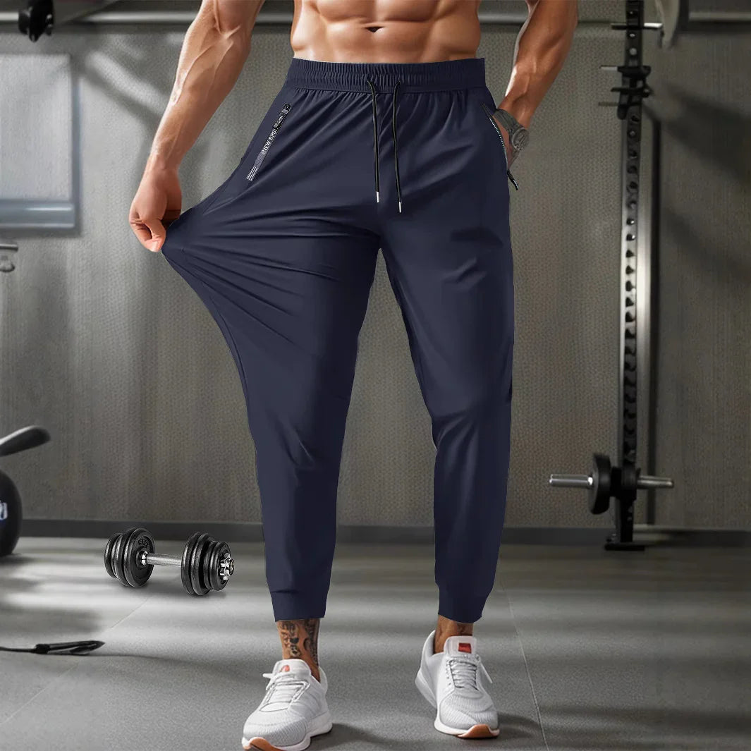 Muscular man stretching navy blue jogger pants in modern gym with dumbbells and workout equipment